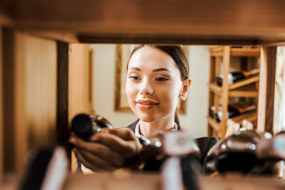 view from shelf at beautiful female wine steward holding bottle at wine store